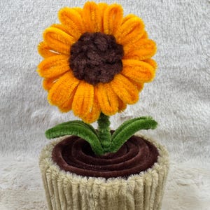 May include: A handcrafted sunflower made of orange and brown pipe cleaners, with green leaves and stem, sits in a tan pot. The flower has a dark brown center and bright orange petals. The pot is a light tan color.