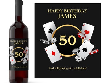 May include: A wine bottle label with a black background and gold accents. The label features playing cards, dice, and poker chips. The text on the label reads "Happy Birthday James 50 And still playing with a full deck!"