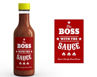 Könnte beinhalten: Ein rotes und weißes Etikett mit dem Text "The Boss With The Sauce" und "Ryan's Slap Yo Mama Recipe". Das Etikett ist an einer Glasflasche mit roter Sauce und gelbem Deckel befestigt.