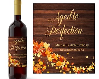 May include: Wine bottle label with a woodgrain background and gold text that reads "Aged to Perfection". The label also features a design of autumn leaves in shades of orange, yellow, and red. The text "Michael's 50th Birthday" and "November 26, 20XX" is also included on the label.