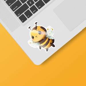 Puede incluir: Una pegatina de abeja de dibujos animados con rayas amarillas y negras, ojos marrones y alas blancas. La abeja sonríe y tiene una expresión linda. La pegatina está en una computadora portátil blanca.