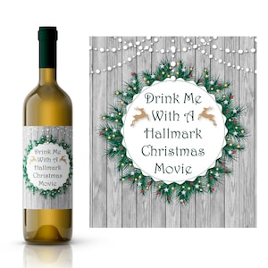 May include: A white wine bottle with a label that says "Drink Me With A Hallmark Christmas Movie". The label features a green wreath with red berries, gold stars, and two gold reindeer. The background is a woodgrain pattern with white lights.