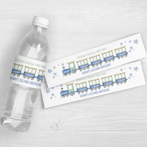 以下が含まれることがあります： 青と緑の電車デザインのラベルが付いた透明なウォーターボトル。「CHUGGA CHUGGA TWO TWO」と「HENRY'S SECOND BIRTHDAY」と書かれています。星のアクセントが付いた、電車をテーマにしたお揃いのラベルも2枚あります。