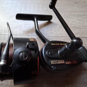 May include: A black Mitchell 300 Pro fishing reel featuring a spool with brown fishing line, a metal handle, and a bail arm. The side of the reel is marked with the text 'ROLLER BEARING' and 'MITCHELL 300 PRO' in gold lettering against the black body.
