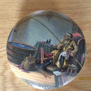 May include: A round, ceramic lid featuring a colourful illustration of a man seated in a chair, holding a fishing rod. The man is wearing a brown coat and a white hat. The scene is set in a room with a wooden floor, a barrel, and a table with various items on it. The text "THE ENTHUSIAST" is printed on the bottom of the lid.