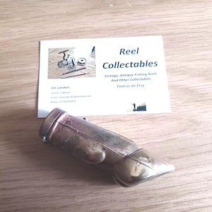 May include: A vintage, brass-toned fishing reel accessory rests on a wooden surface, next to a business card. The card reads "Reel Collectables" and lists contact information. The accessory has a rounded body and a pointed end.
