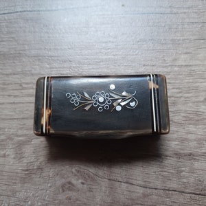 Puede incluir: Una caja rectangular de color marrón oscuro con un diseño floral decorativo con incrustaciones de detalles blancos y dorados. La caja tiene un borde similar al carey y probablemente se utiliza para guardar objetos pequeños. El diseño está centrado en la tapa.