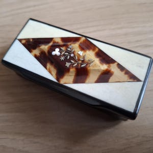 May include: A rectangular box with a lid featuring a central diamond-shaped inlay of brown and black tortoiseshell pattern. The inlay is decorated with a delicate silver floral motif. The surrounding lid surface is a smooth, off-white material.