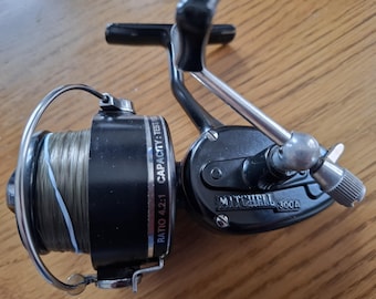 May include: A black Mitchell 300A fishing reel with a silver handle and spool. The reel has a ratio of 4.2:1 and the text "CAPACITY: TEST" is visible. The spool is filled with fishing line.