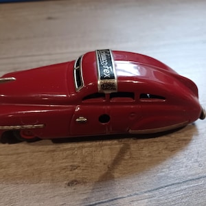 May include: Red toy car with a silver trim and a black and silver "Schuco-Tex" logo on the roof.