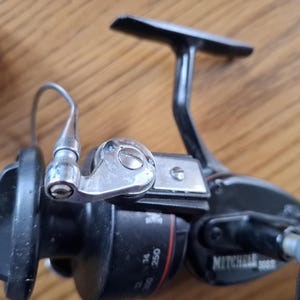 May include: A black and silver Mitchell 300S fishing reel. The reel has a handle, a spool, and a line guide. The reel is designed for freshwater fishing. The reel is made of metal and plastic.