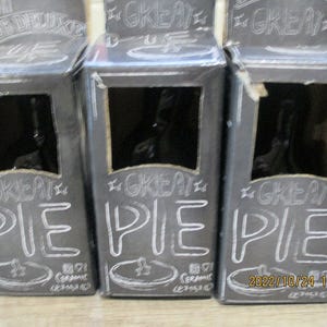 Op de afbeelding: Drie zwarte en witte kartonnen dozen met de woorden "Great Pie" in wit op de voorkant. Elke doos heeft een klein venster dat een zwarte keramische taartvorm in de doos laat zien.
