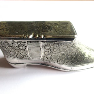 Puede incluir: Una caja de alhajas de plata con forma de zapato con un diseño grabado ornamentado. La tapa está articulada y tiene un patrón floral.
