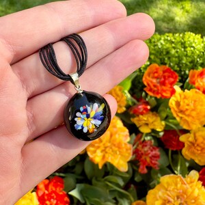 May include: A black glass pendant necklace with a colorful flower design. The pendant is round and has a silver bail. The necklace is on a black cord.