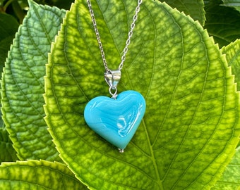 May include: A silver chain necklace with a light blue heart-shaped pendant. The pendant has a swirl pattern and is hanging against a green leaf background.
