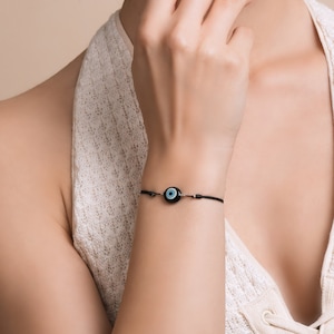 Op de afbeelding: Een zwart koordarmband met een blauwe kwade oog bedel. De bedel is rond en heeft een wit centrum.