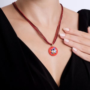 May include: A red glass pendant with a floral design hangs from a multi-strand necklace. The necklace is made of thin, red cords.
