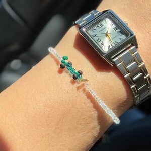 May include: A silver watch with a square face and Roman numeral markings. The watch is on a person's wrist with a beaded bracelet with green and white beads.