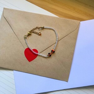 May include: A delicate beaded bracelet with a gold clasp and small orange, black, and white beads. The bracelet is lying on a brown envelope with a red heart sticker.