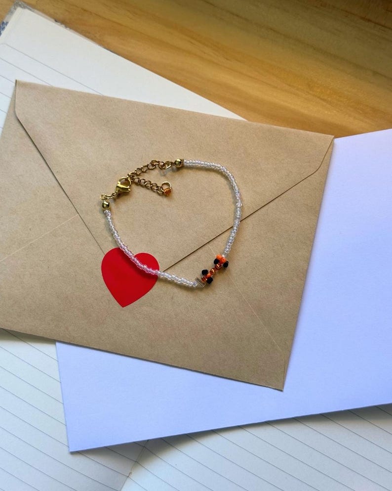 May include: A delicate beaded bracelet with a gold clasp and small orange, black, and white beads. The bracelet is lying on a brown envelope with a red heart sticker.