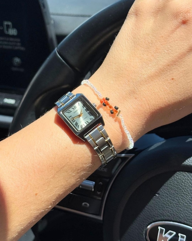 May include: A silver and gold toned watch with a square face and Roman numeral markings. The watch is on a person's wrist with a white beaded bracelet with orange and black beads.