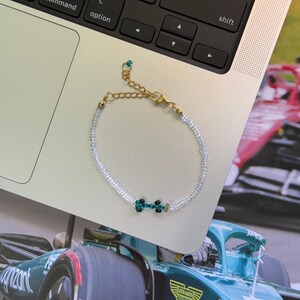 May include: A white beaded bracelet with a green and black charm in the shape of a race car. The bracelet has a gold clasp.
