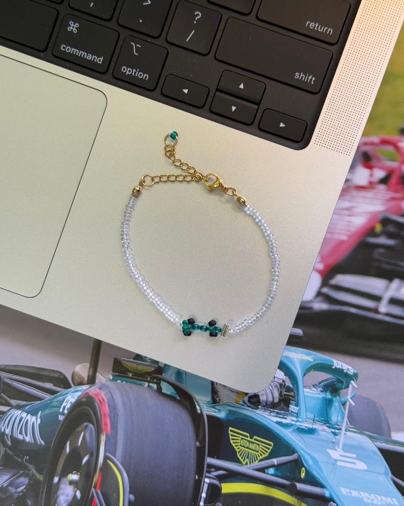 May include: A white beaded bracelet with a green and black charm in the shape of a race car. The bracelet has a gold clasp.