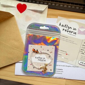 May include: A white beaded bracelet with a small orange and black charm. The bracelet is on a white card with the text "kaitlyn in reverie" and a cloud design. The card is in a clear iridescent bag.