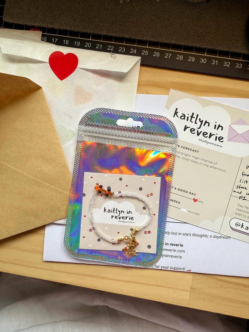 May include: A white beaded bracelet with a small orange and black charm. The bracelet is on a white card with the text "kaitlyn in reverie" and a cloud design. The card is in a clear iridescent bag.