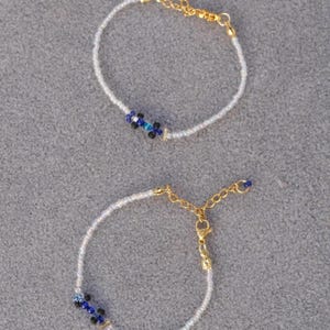 May include: Two delicate beaded bracelets. Each bracelet features small white beads with a cluster of blue and black beads at the center. Gold-colored chain and clasp complete the design. The bracelets are displayed on a gray surface.