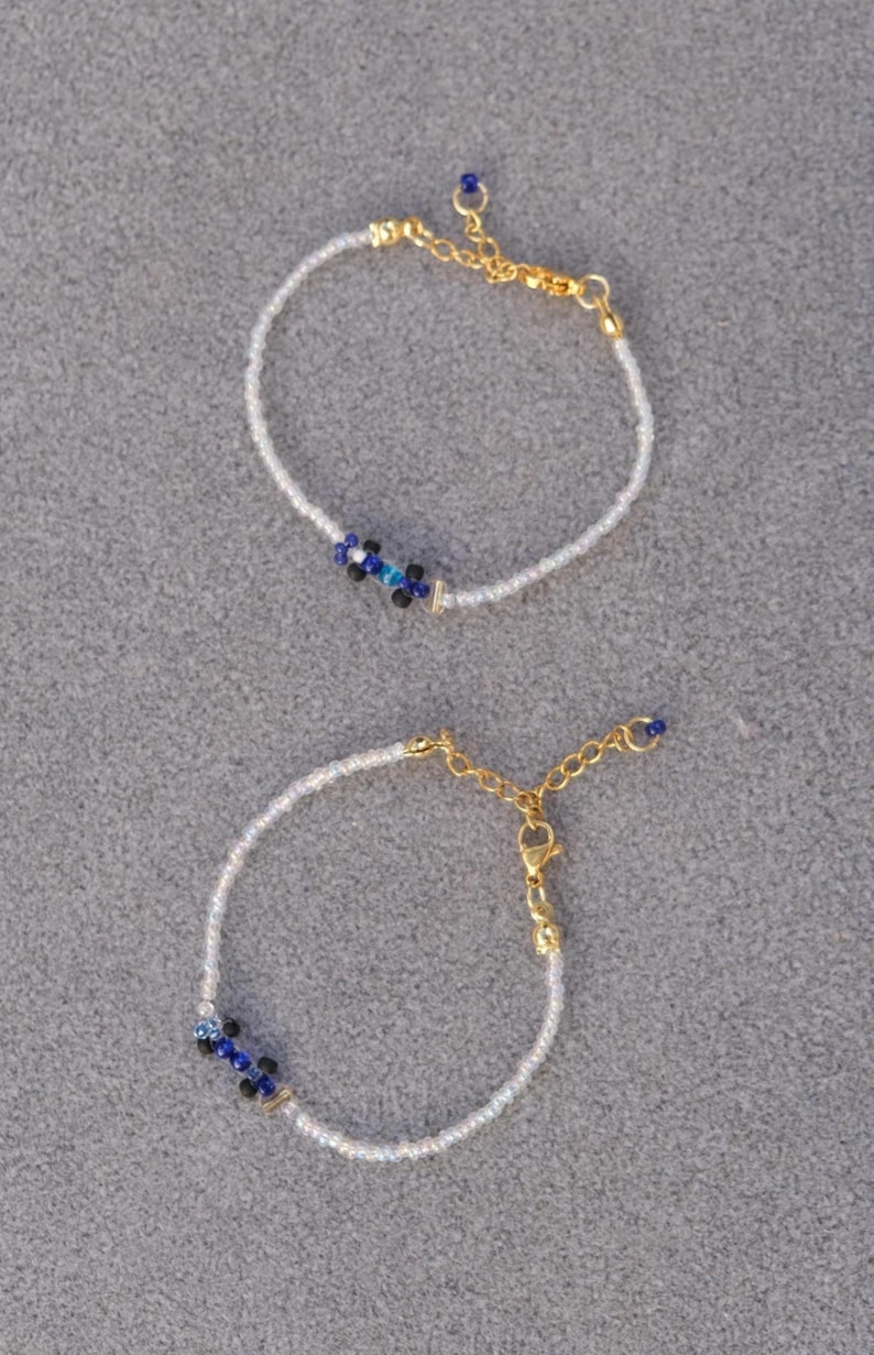 May include: Two delicate beaded bracelets. Each bracelet features small white beads with a cluster of blue and black beads at the center. Gold-colored chain and clasp complete the design. The bracelets are displayed on a gray surface.