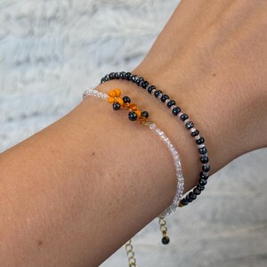 May include: Two beaded bracelets on a wrist. One bracelet is made of small black beads and the other is made of small white beads. A small orange and black beaded charm hangs from the white bracelet.