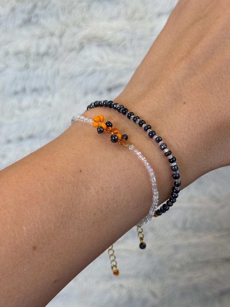 May include: Two beaded bracelets on a wrist. One bracelet is made of small black beads and the other is made of small white beads. A small orange and black beaded charm hangs from the white bracelet.