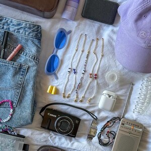 May include: An assortment of accessories including a brown purse, blue sunglasses, a purple cap, and a Canon digital camera. Also visible are beaded eyeglass chains, a wristwatch, a small radio, and a pair of sandals. The items are arranged on a white surface.