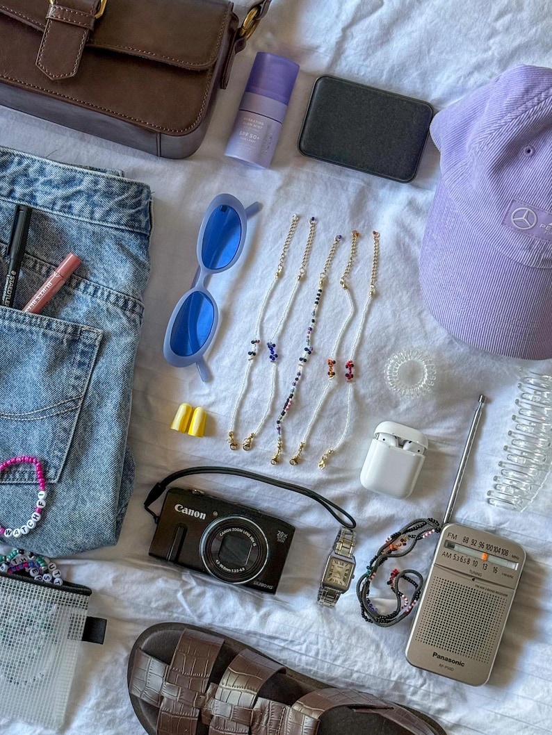 May include: An assortment of accessories including a brown purse, blue sunglasses, a purple cap, and a Canon digital camera. Also visible are beaded eyeglass chains, a wristwatch, a small radio, and a pair of sandals. The items are arranged on a white surface.