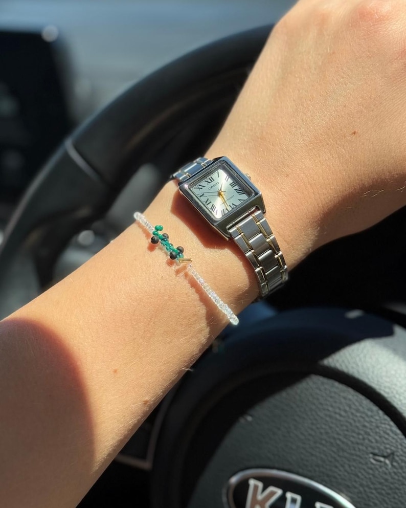 May include: A silver watch with a square face and Roman numeral markings. The watch is on a person's wrist with a beaded bracelet with green and white beads.