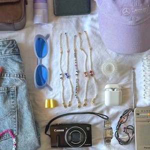 May include: A flat lay of various items including a brown leather purse, a purple hat, a camera, a pair of sunglasses, a pair of earplugs, a small white box, a radio, a watch, a pair of beaded bracelets, a pair of beaded necklaces, a pair of hair ties, a bottle of hand sanitizer, a small black box, and a pair of jeans.