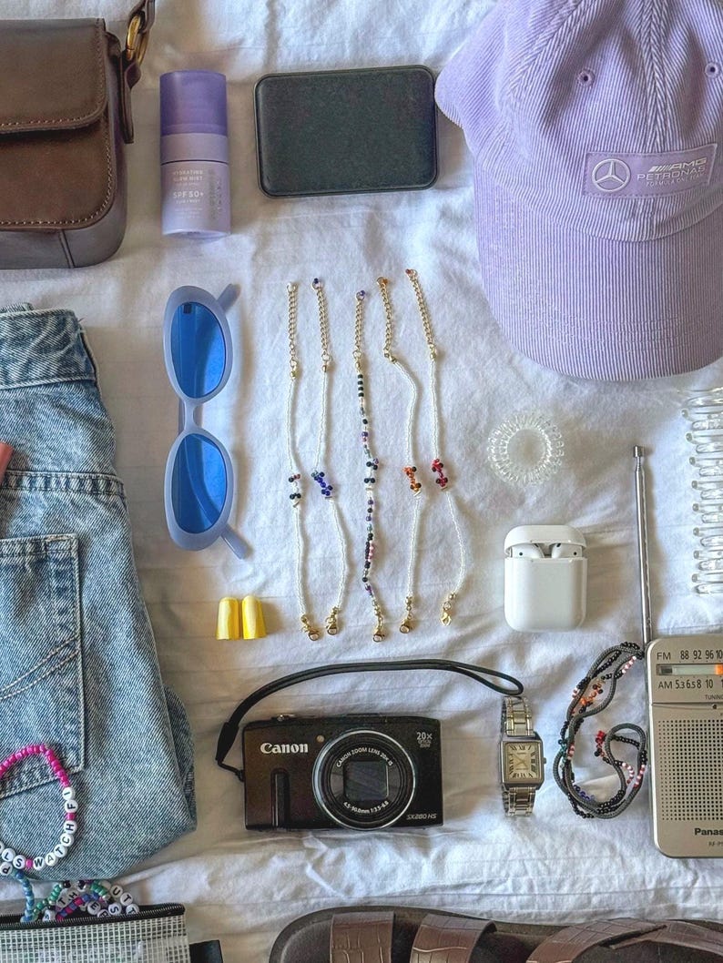 May include: A flat lay of various items including a brown leather purse, a purple hat, a camera, a pair of sunglasses, a pair of earplugs, a small white box, a radio, a watch, a pair of beaded bracelets, a pair of beaded necklaces, a pair of hair ties, a bottle of hand sanitizer, a small black box, and a pair of jeans.