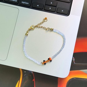 May include: A delicate beaded bracelet with a small orange and black car charm. The bracelet is made of tiny white beads and has a gold clasp.