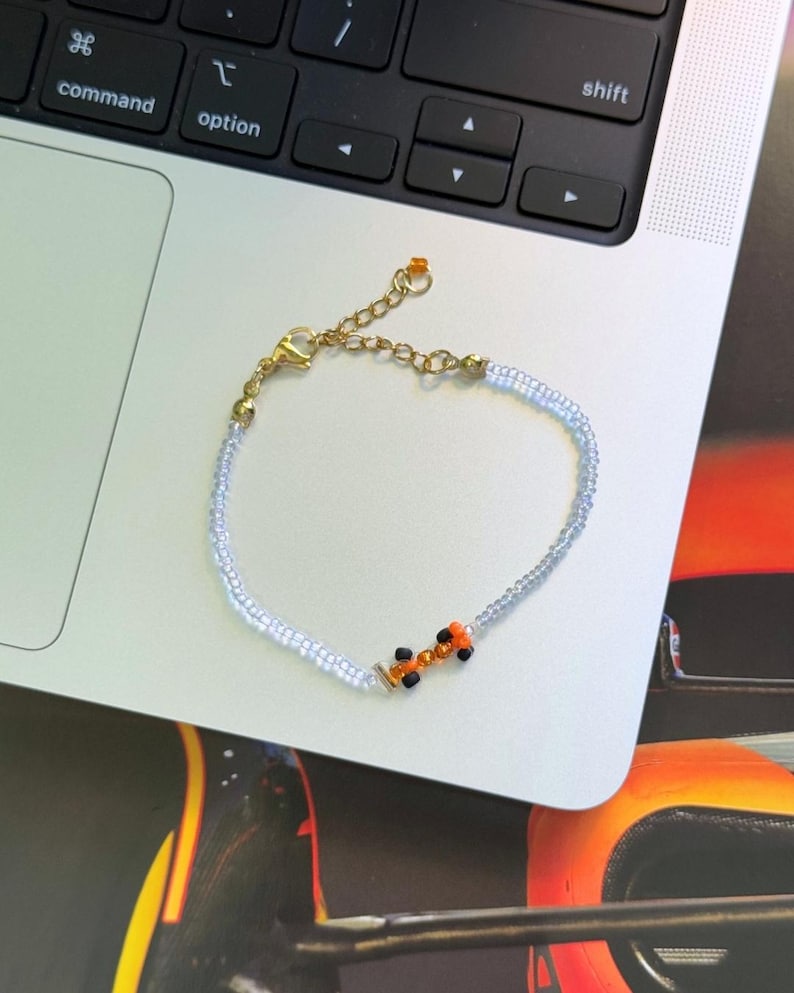 May include: A delicate beaded bracelet with a small orange and black car charm. The bracelet is made of tiny white beads and has a gold clasp.
