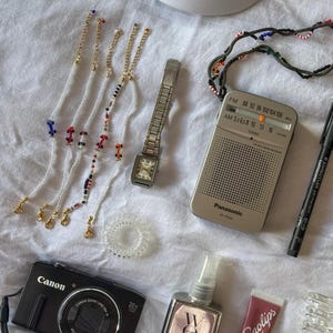 May include: A flat lay of various items including a black camera, a white baseball cap with a black logo, a silver radio with the text "Panasonic RF-P100", a silver watch, a clear hair scrunchie, a pink lip balm tube with the text "Lanolips", a clear bottle with a pink label and the text "WOW DREAM COAT", and several beaded bracelets with gold clasps.