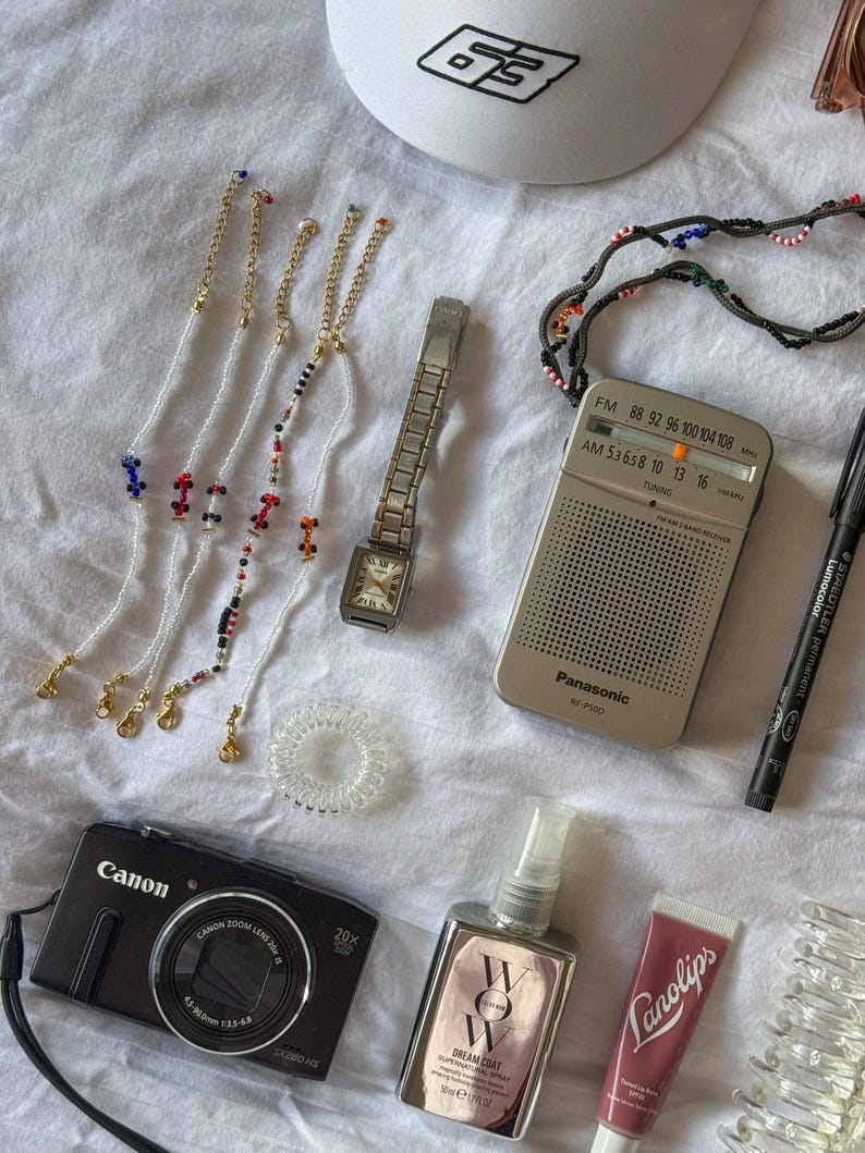 May include: A flat lay of various items including a black camera, a white baseball cap with a black logo, a silver radio with the text "Panasonic RF-P100", a silver watch, a clear hair scrunchie, a pink lip balm tube with the text "Lanolips", a clear bottle with a pink label and the text "WOW DREAM COAT", and several beaded bracelets with gold clasps.