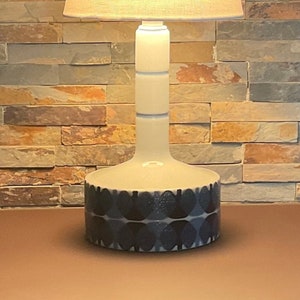 Può includere: Una lampada da tavolo in ceramica bianca con un paralume in tessuto bianco. La base della lampada ha un motivo geometrico blu e bianco.