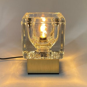 May include: A clear glass table lamp with a square base and a gold metal base. The lamp is lit and the light shines through the glass.