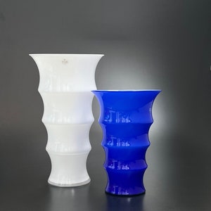 May include: Two glass vases, one white and one blue, with a textured, ribbed design. The white vase has the text "Kosta Boda" on the side.