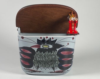 May include: A ceramic container with a blue and white floral design and a wooden lid. A small red figurine is attached to the lid.