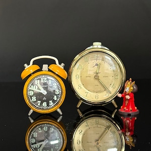 Könnte beinhalten: Zwei Wecker, einer gelb und einer goldfarben, mit schwarzen Zifferblättern. Der gelbe Wecker hat ein weißes Zifferblatt mit schwarzen Zahlen und Zeigern. Der goldene Wecker hat ein weißes Zifferblatt mit schwarzen römischen Ziffern und Zeigern. Eine kleine rote Figur steht vor dem goldenen Wecker.