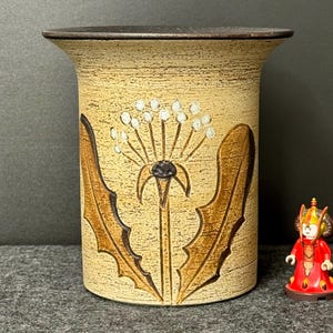 May include: A beige ceramic vase with a dandelion design in brown and white. The vase has a cylindrical shape with a slightly flared top. A small red figurine is next to the vase.