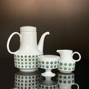 May include: A white ceramic coffee pot, creamer, and sugar bowl with a geometric pattern in shades of green, blue, and white. The set is on a black surface.