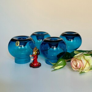 Può includere: Quattro portacandele in vetro blu turchese con forma arrotondata. I portacandele sono disposti a semicerchio su una superficie bianca.